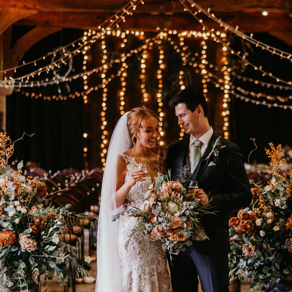 Bride in lace strap wedding dress with tulle skirt and veil holding large orange and green bridal bouquet stands holding a glass of sparkling wine with groom in dark suit and blue tie after rustic wedding ceremony