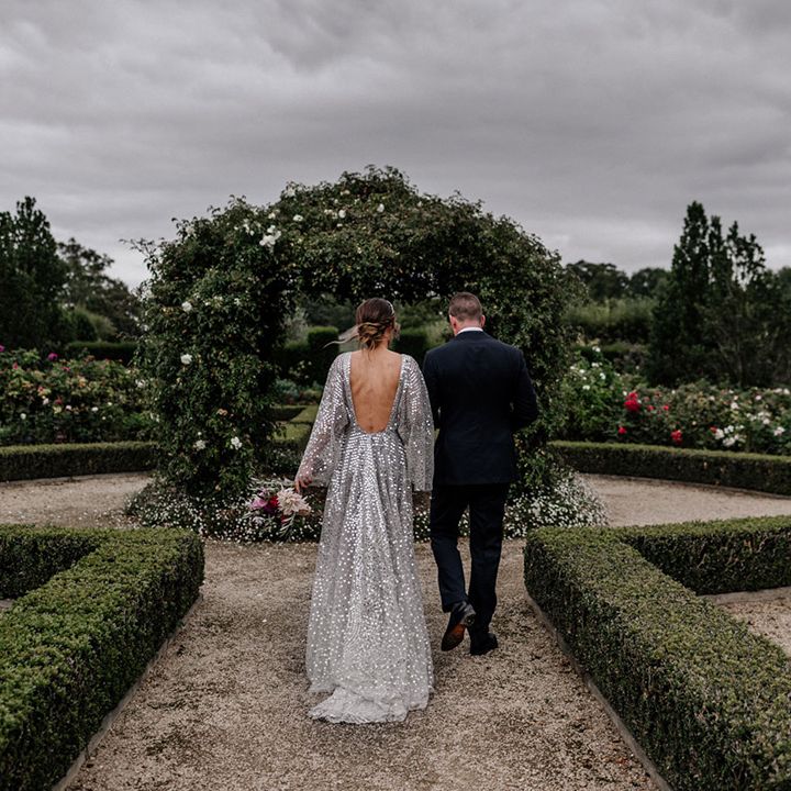 Bride in a low back silver sparkly wedding dress and groom in a navy suit arm in arm in their garden wedding 