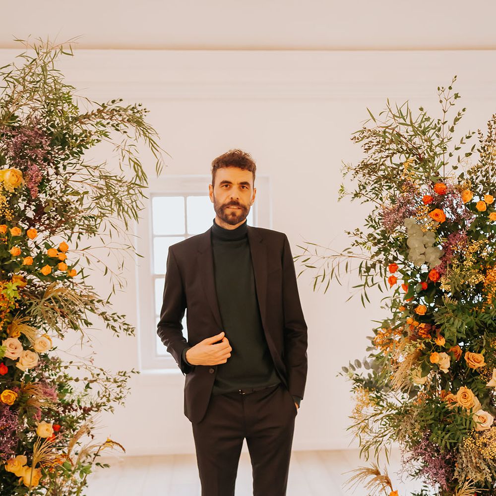 Groom in an all black suit and top standing in between orange, yellow and foliage column wedding flower arrangement 