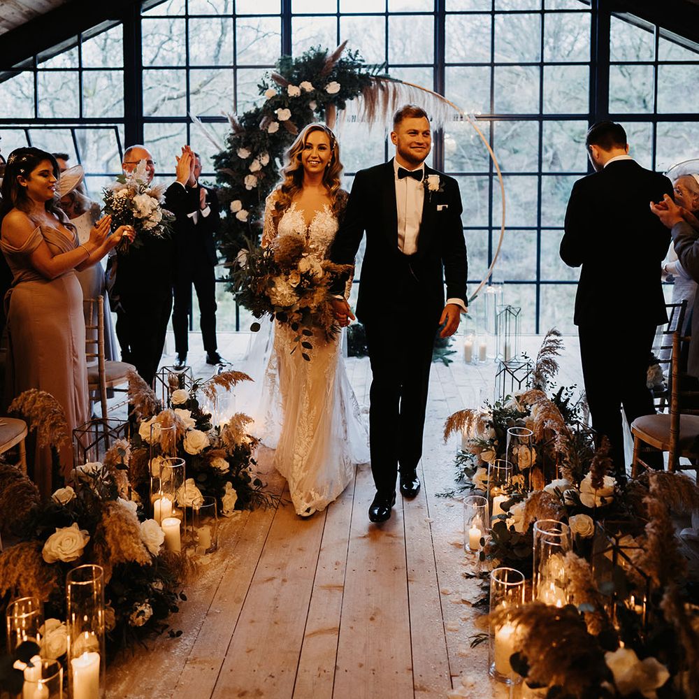 Bride in lace wedding dress and groom in black tie walk back down the aisle as a married couple with floral moongate at the altar