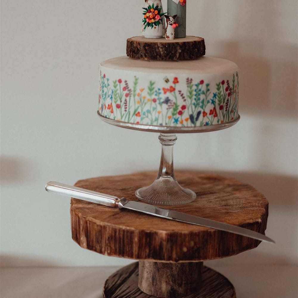 Wedding cake with white frosting with colourful flowers and bees with custom wooden cake toppers of bride, groom and their cat