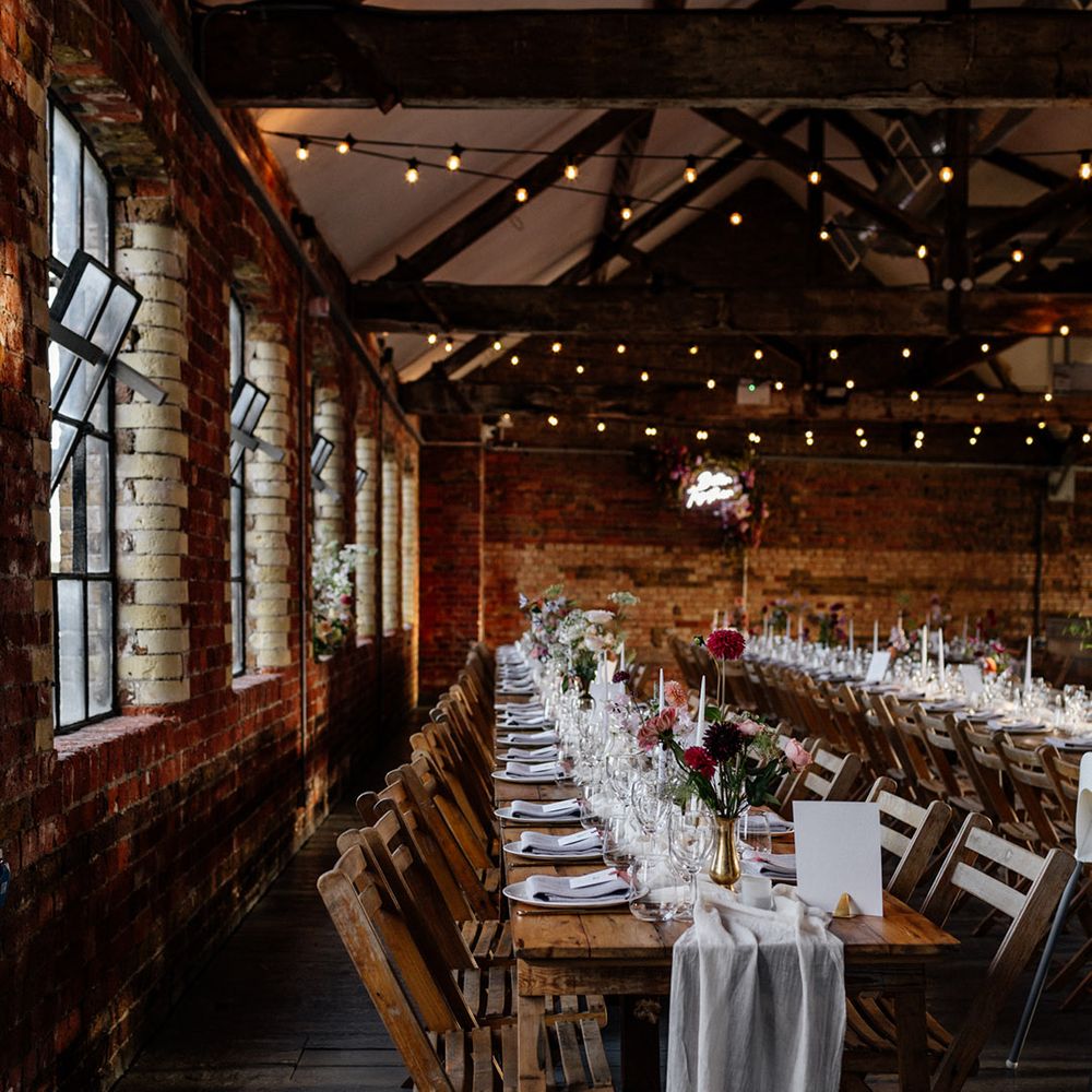 Industrial wedding venue with wooden banquet tables decorated with fabric table runners and gold vessels filled with flower stems 