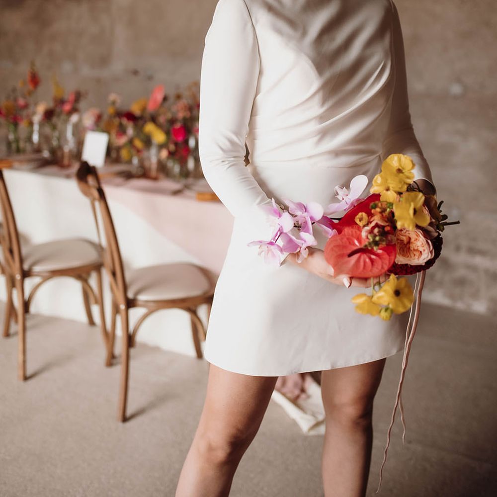 Bride in a short wedding dress with long sleeves holding a pink anthurium, purple orchis and yellow flower wedding bouquet 