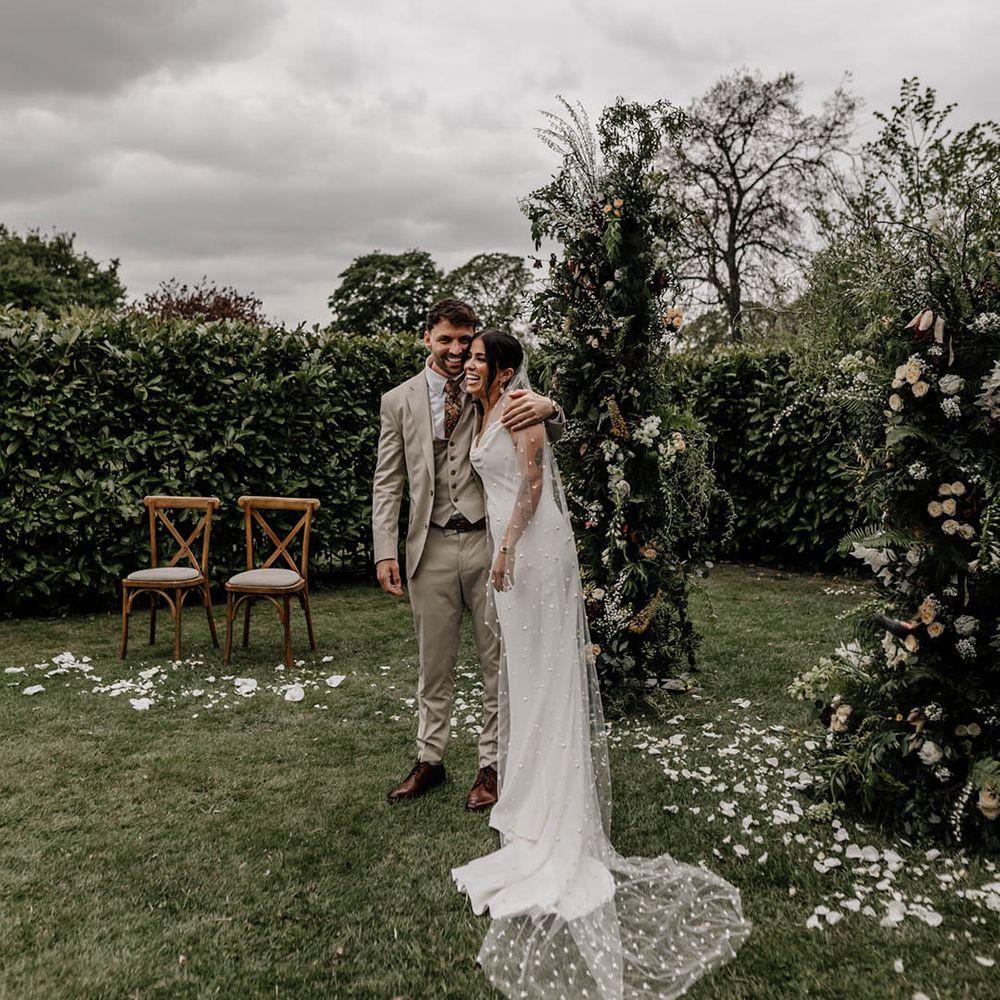 Bride in a minimalist slip wedding dress and bespoke pearl veil at her outdoor wedding ceremony with greenery column flowers