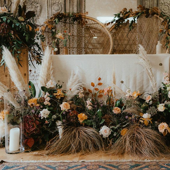 Boho altar setting with peacock chairs, pampas grass, orange wedding flowers and candles in hurricane vases 