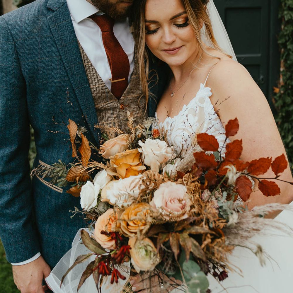 Groom in a blue suit with a brown waistcoat and burnt orange tie with gold tie clip with the bride as she leans against him carrying an autumnal bouquet in sequin Blush by Hayley Paige wedding dress