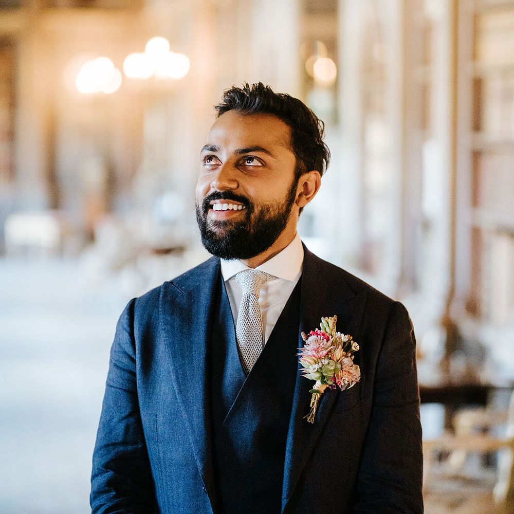 Groom wears three piece blue suit and grey textured tie with floral buttonhole 