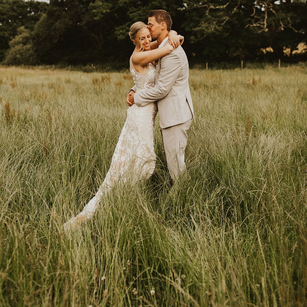 Groom in a light wedding suit hugs the bride as he kisses her on the forehead for their couple portraits 