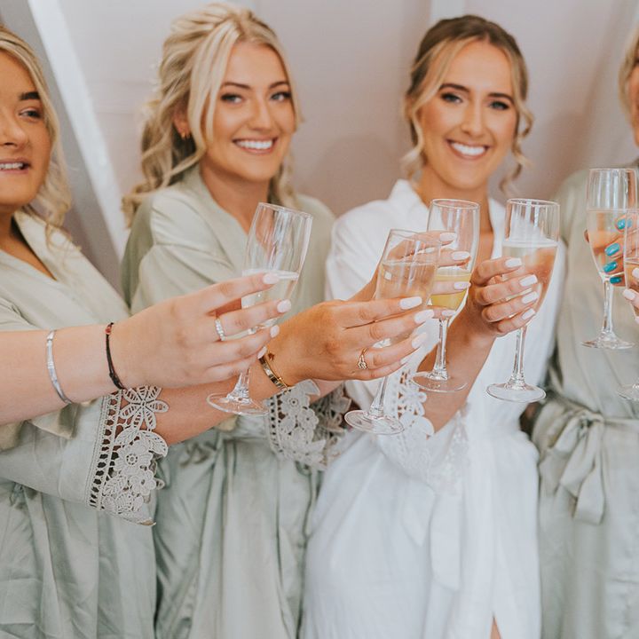 Bride in a white satin robe with the bridesmaids in sage green bridesmaid robes all drinking some champagne