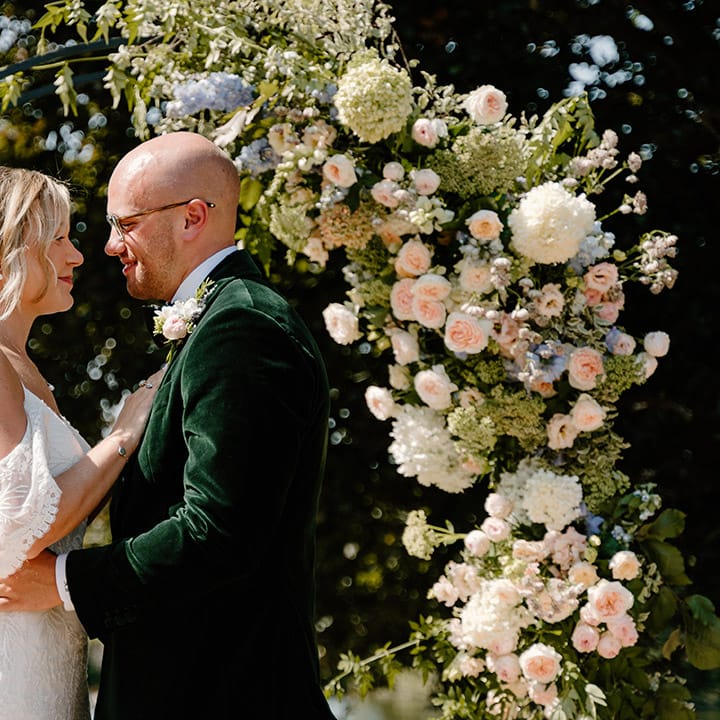 Bride wears lace off the shoulder wedding dress whilst stood with her groom in green velvet black tie 