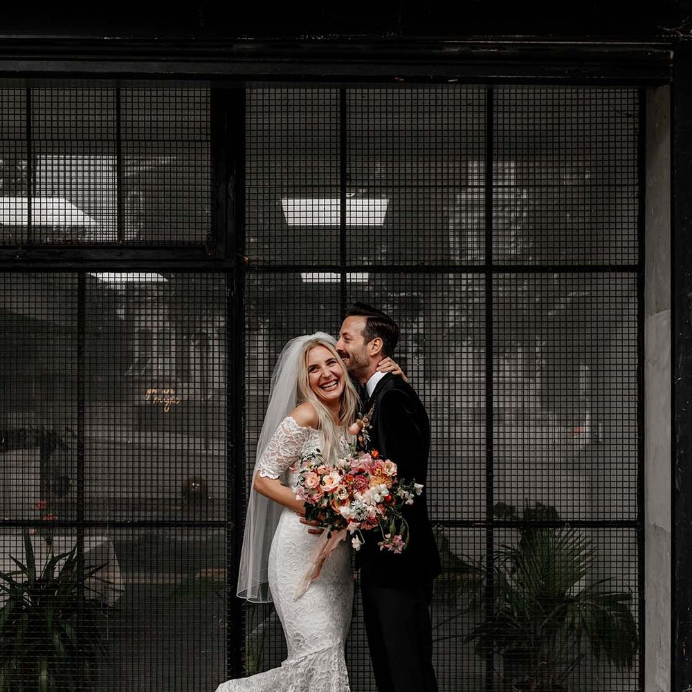 Bride in boho two piece lace bridal outfit with large colourful mixed bouquet and groom in black tux with velvet look bow tie embracing at Hackney Studios