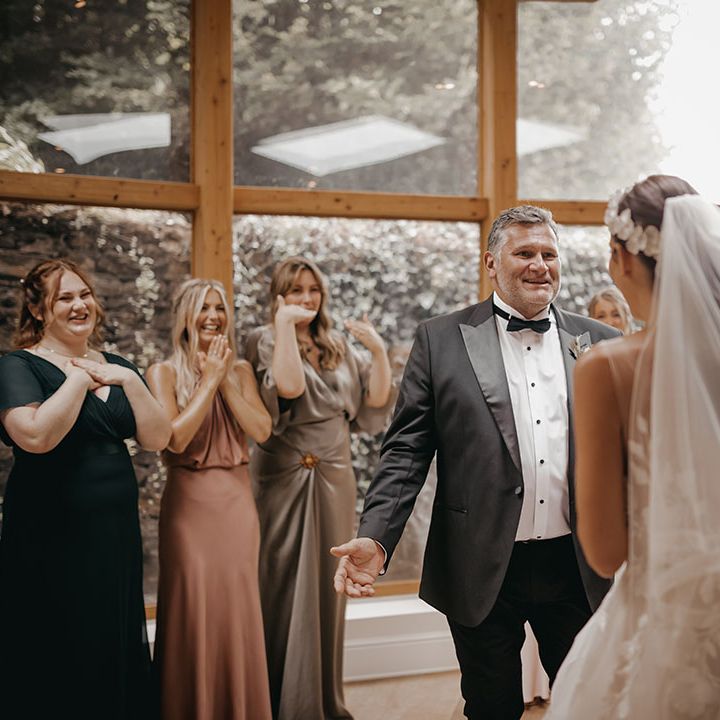 Father of the bride in a black tuxedo and bridesmaids gets their first look at the bride