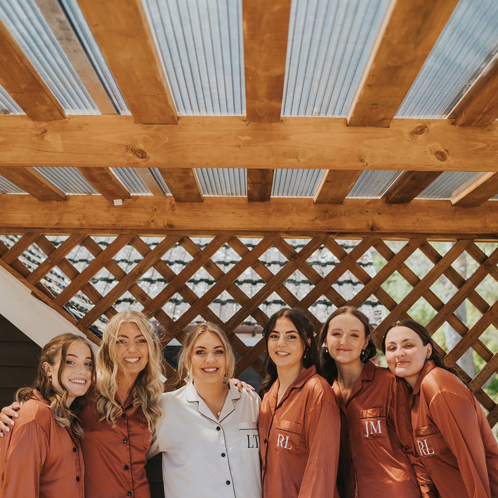 Bride in white pyjamas stands with her bridesmaids who wear matching pyjamas in burnt orange and personalised lettering 