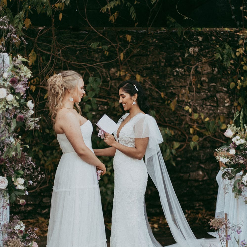 Brides in sleeveless ruched wedding dress and short sleeve v neck wedding dress with puff lace shoulders, reading vows to each other and matching earrings