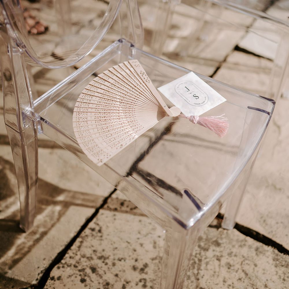 Clear acrylic chairs with biodegradable confetti and wooden fans for guests during the summer weather 