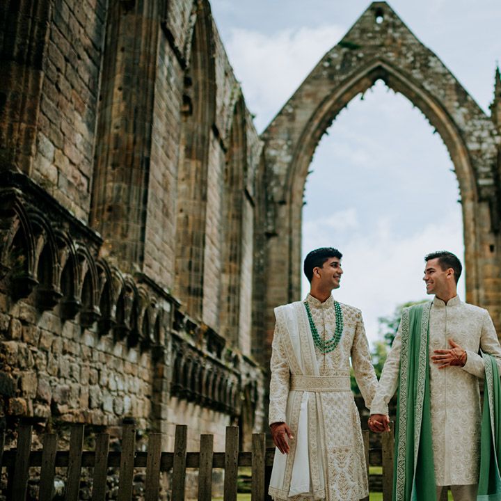 Grooms look lovingly at one another whilst holding hands wearing matching sherwani's outdoors for couples portraits