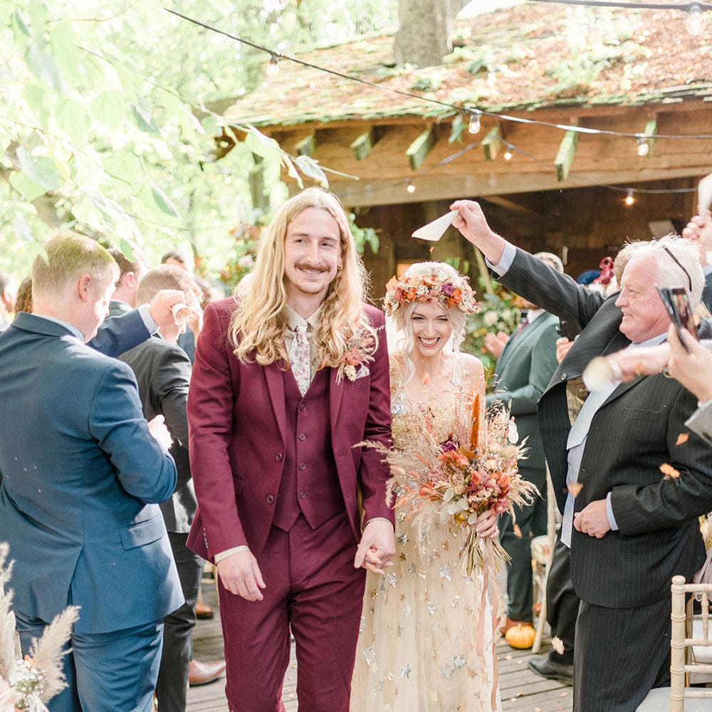 Groom in maroon suit with floral tie and matching pocket square walks alongside his bride in embellished wedding dress with sequin stars