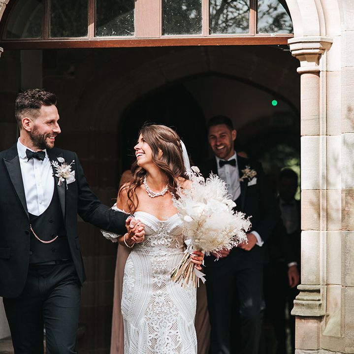 Groom in black tuxedo with gold chain with bride in fitted lace wedding dress and detachable sleeves walking out of the ceremony carrying a dried flower bouquet
