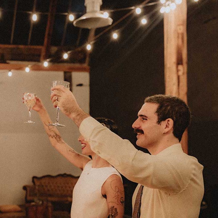 Lisa & Gabriel lift their glasses in cheers during industrial styled reception with festoon lighting 