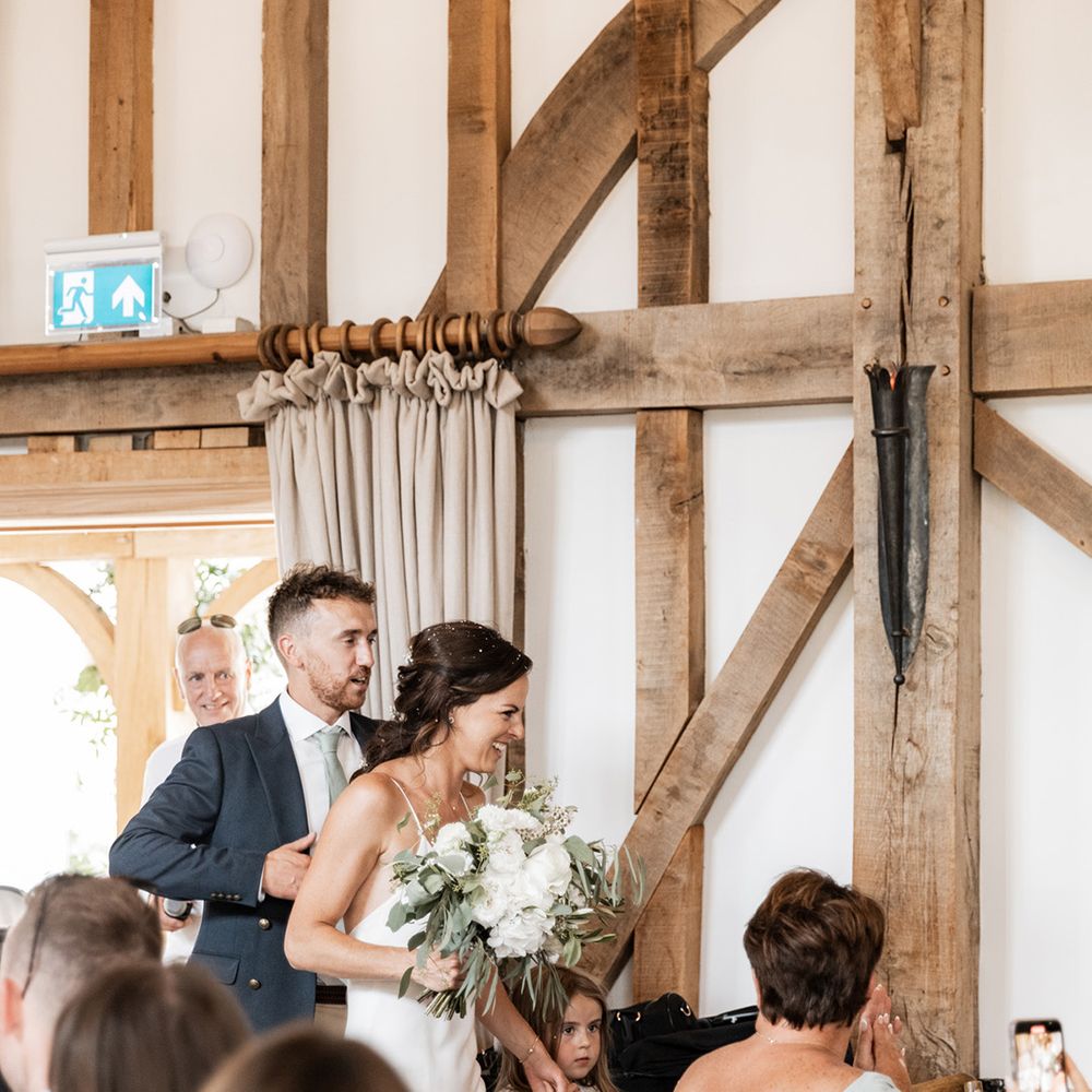 Bride carries white floral bouquet and walks into rustic barn wedding venue for reception 
