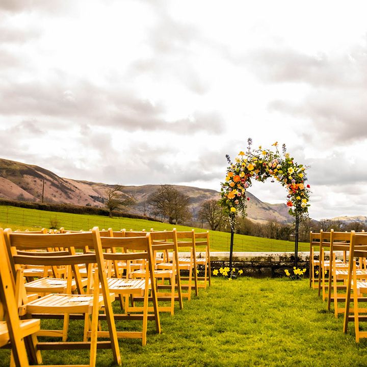 Colourful floral archway to the front of outdoor aisle lined by wooden chairs 