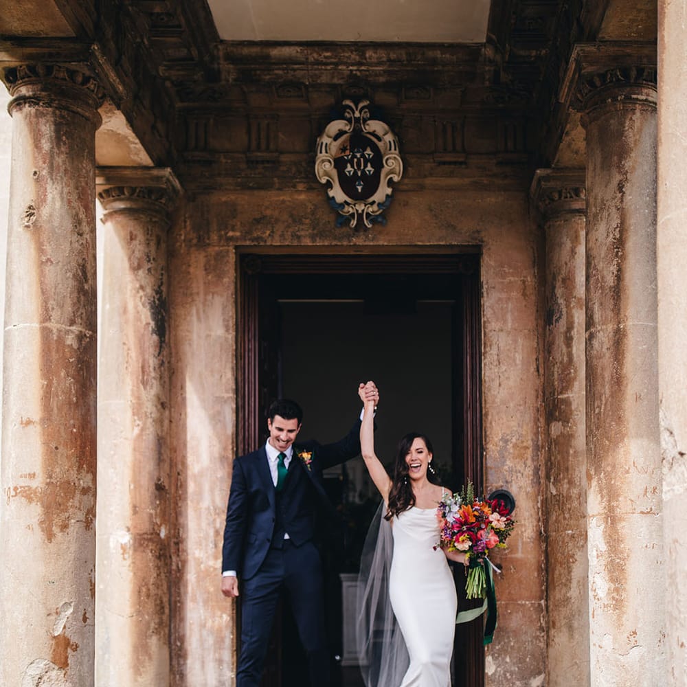Bride in white satin cowl neck Kyha wedding dress exiting Elmore Court with the groom in a three piece navy suit and green tie 