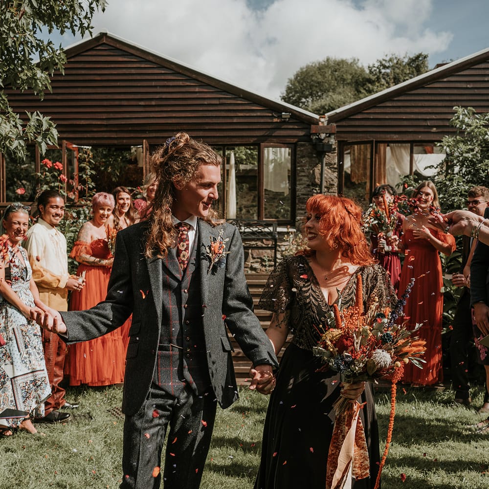 Bride in embellished black wedding dress holding large orange dried flower and eucalyptus bouquet with orange ribbon holding hands with groom in dark grey three piece suit and burgundy flower patterned tie doing orange confetti exit 