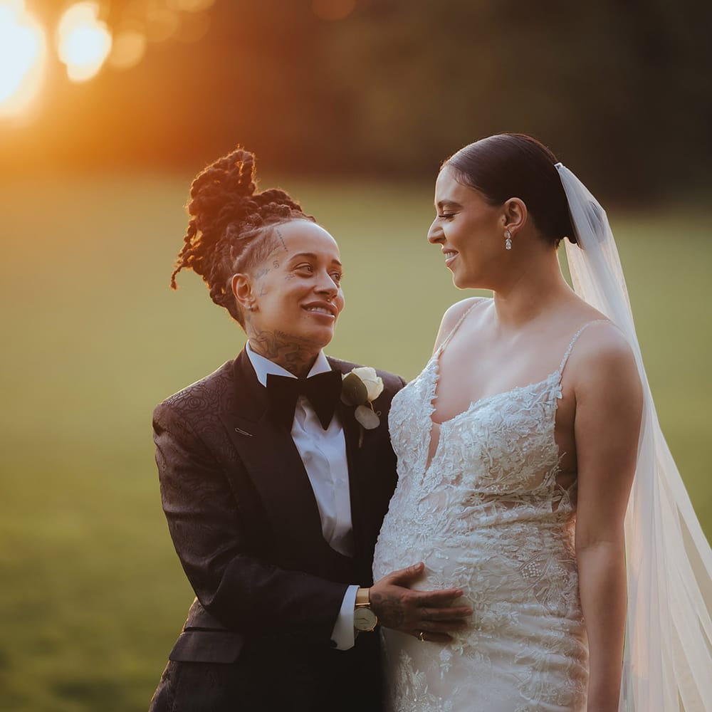 Bride in black snakeskin print tuxedo with black bowtie and white rose boutonniere and golden watch holding bride's baby bump, wearing lace sleeveless v neck Enzoani wedding dress with tulle finish and church length veil at golden hour couples photoshoot 