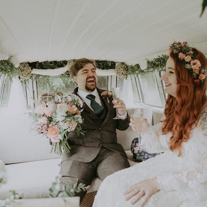 The groom in a three piece suit with the bride in a boho wedding dress with flower crown ride in the VW camper van wedding transport decorated with flowers