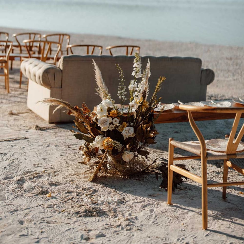 White sofa and wooden chairs on beach wedding with pampas grass, burnt orange dried flower, white garden rose and peony flower arrangements 
