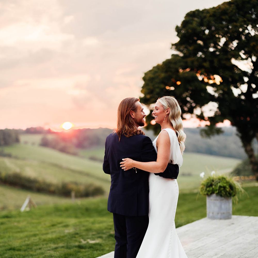 Gorgeous sunset wedding photography with the bride in a draped back wedding dress with a train embracing the groom in a blue suit 