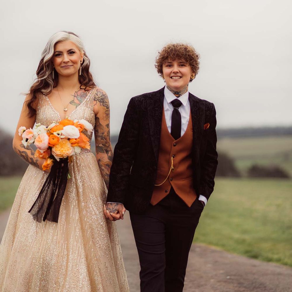 Bride in sleeveless deep v neck sparkly gold wedding dress holding orange garden rose, white poppy, light pink carnation and foliage bridal bouquet wrapped in sparkly gold ribbon with bride in dark velvet bridal jacket with rust waistcoat and pocket square, black suit trousers and gold pocket watch at Cotswolds wedding venue