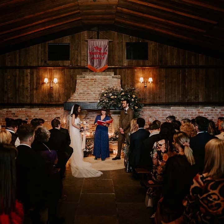 Groom in chocolate brown grooms suit standing with bride in strapless ivory fringed crepe gown and chapel length veil and celebrant in deep blue playsuit standing by large fireplace at Willow Marsh Farm Loughborough 