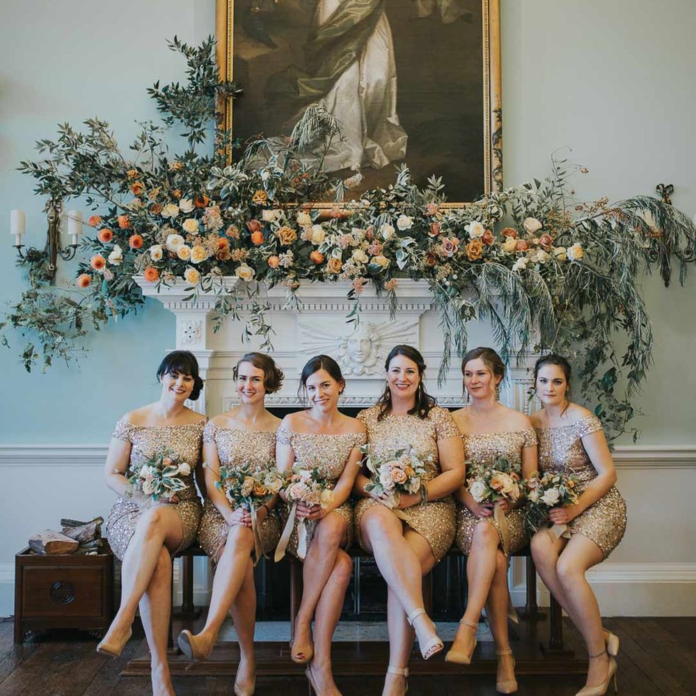 Bridesmaids in off shoulder short golden bridesmaid dresses sitting underneath large portrait at Oxfordshire wedding venue