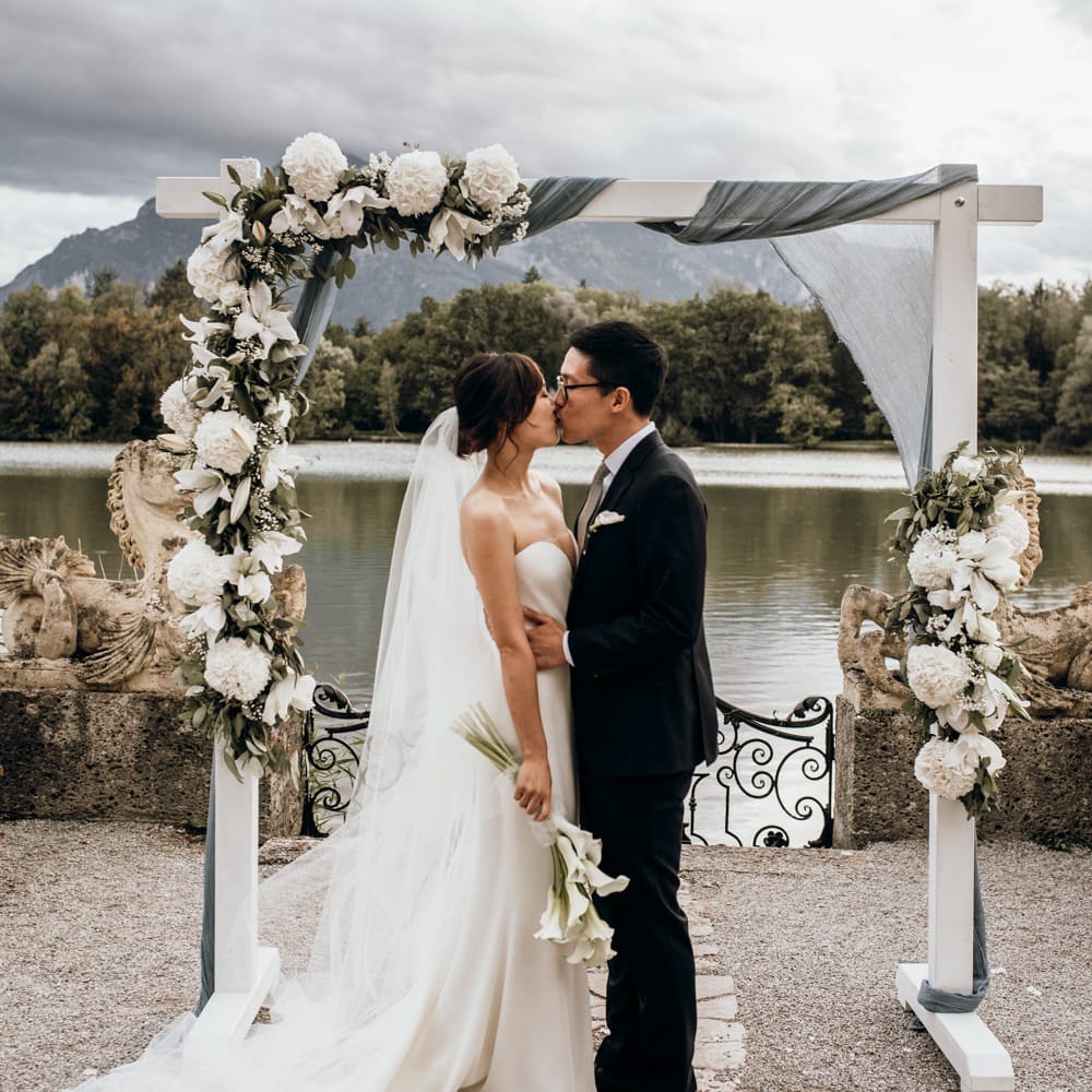 Bride and groom kissing next to a wooden frame decorated with drapes and white flowers 