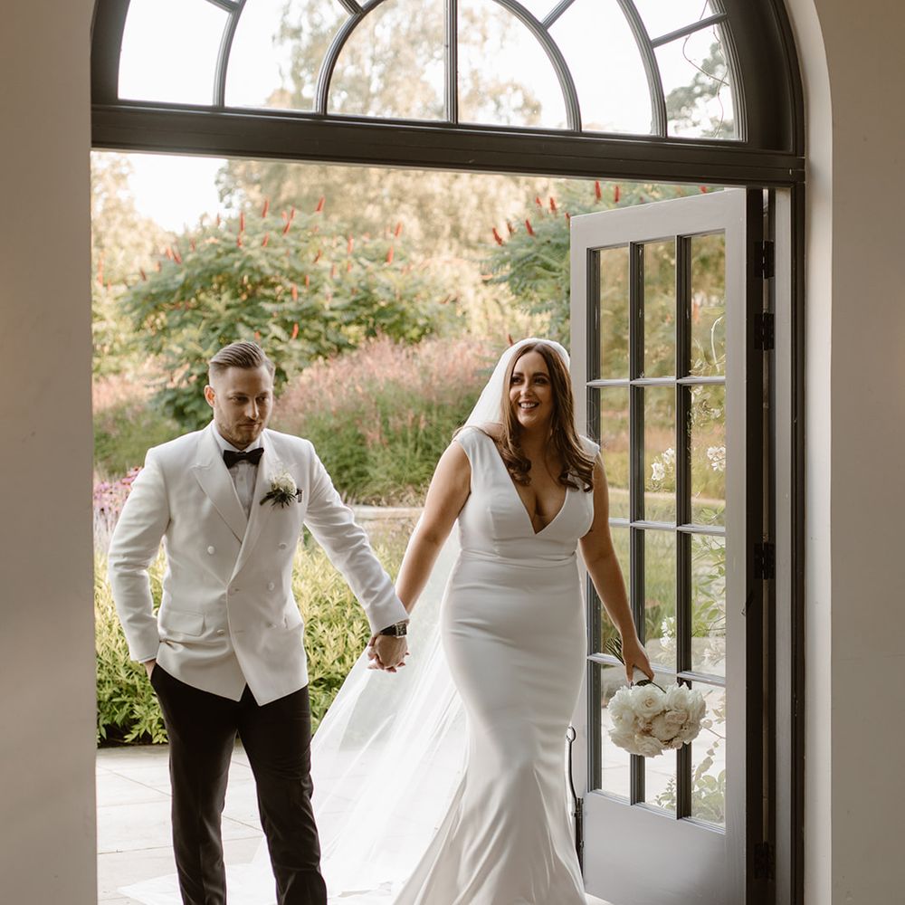 Bride in Made With Love wedding dress and groom in white double breasted tuxedo jacket enter the Fig House at Middleton Lodge holding hands