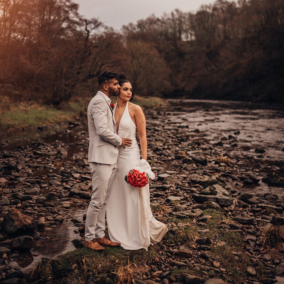 Bride & groom kiss outdoors as bride holds red floral bouquet 