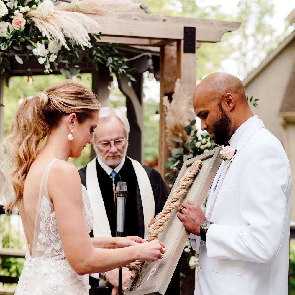Bride in white Made With Love embroidered wedding dress and curled ponytail ties knot held by groom in white tuxedo jacket during wedding ceremony at backyard wedding