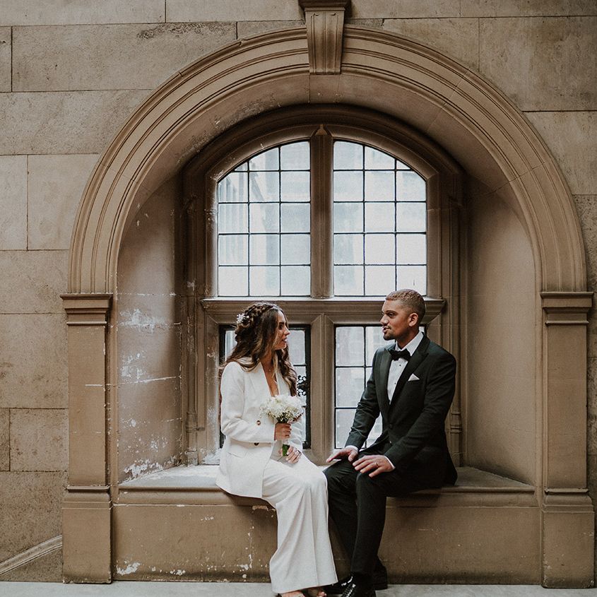 Groom in a tuxedo and bride in a white suit sitting on a window ledge at their Sheffield registry office wedding 