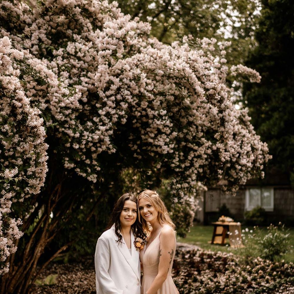Two brides in a white wedding suit and blush wedding dress embracing under a blossom tree
