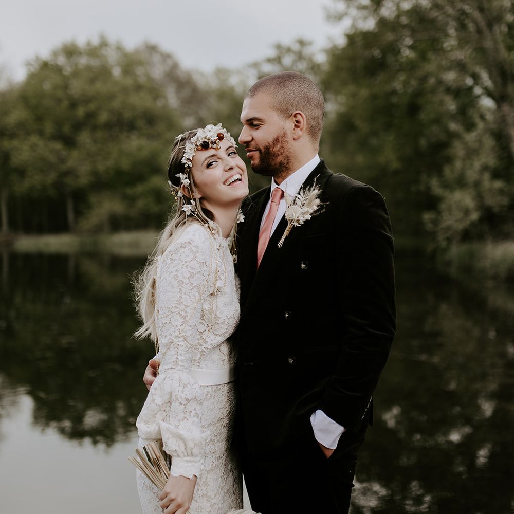 Groom in a black suit with corduroy jacket embracing his boho bride in a lace wedding dress as she holds a dried palm leaf and flower wedding bouquet 