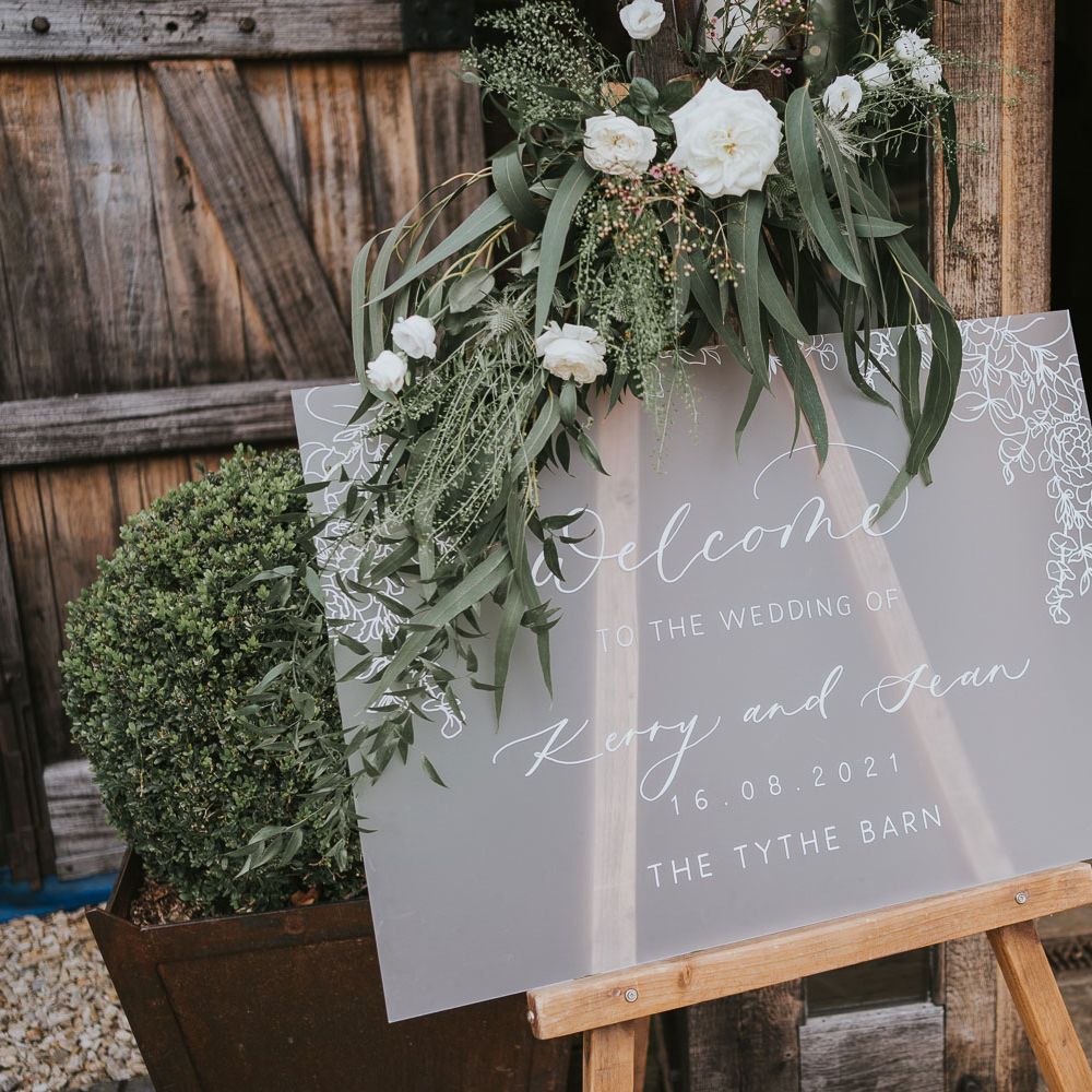 Frosted acrylic wedding welcome sign with leaf details topped with white floral decor at Tythe Barn wedding with barn wedding flowers