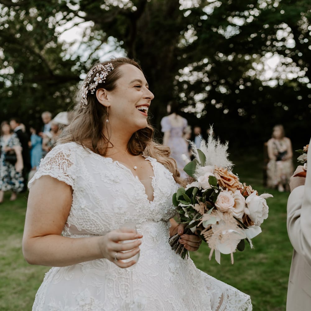 Birde in homemade lace wedding dress with pearl and floral hair pin holding mixed rose bouquet smiles at groom in beige suit at outdoor wedding reception