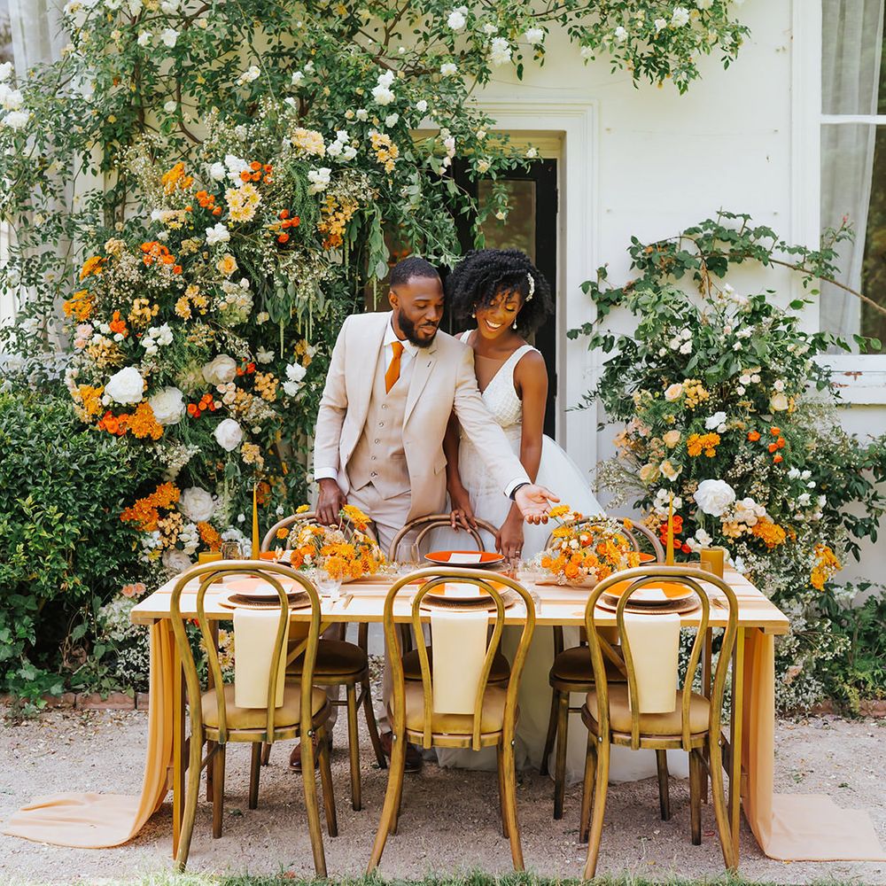 Black bride and groom in a beige suit and plunging neckline wedding dress at their outdoor wedding reception with orange decor and flowers 