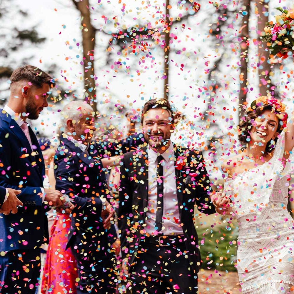 Bride in off the shoulder Grace Loves Lace wedding dress and bridal flower crown holds colourful wedding bouquet and the hand of groom in black suit as guests throw multicoloured confetti during outdoor ceremony at The Bridal Barn wedding venue