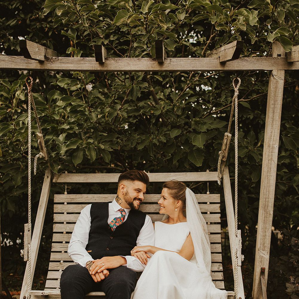 Bride and groom portrait sitting on a wooden swing with groom in a waistcoat and floral tie and bride in bow front sandals and cathedral length veil 
