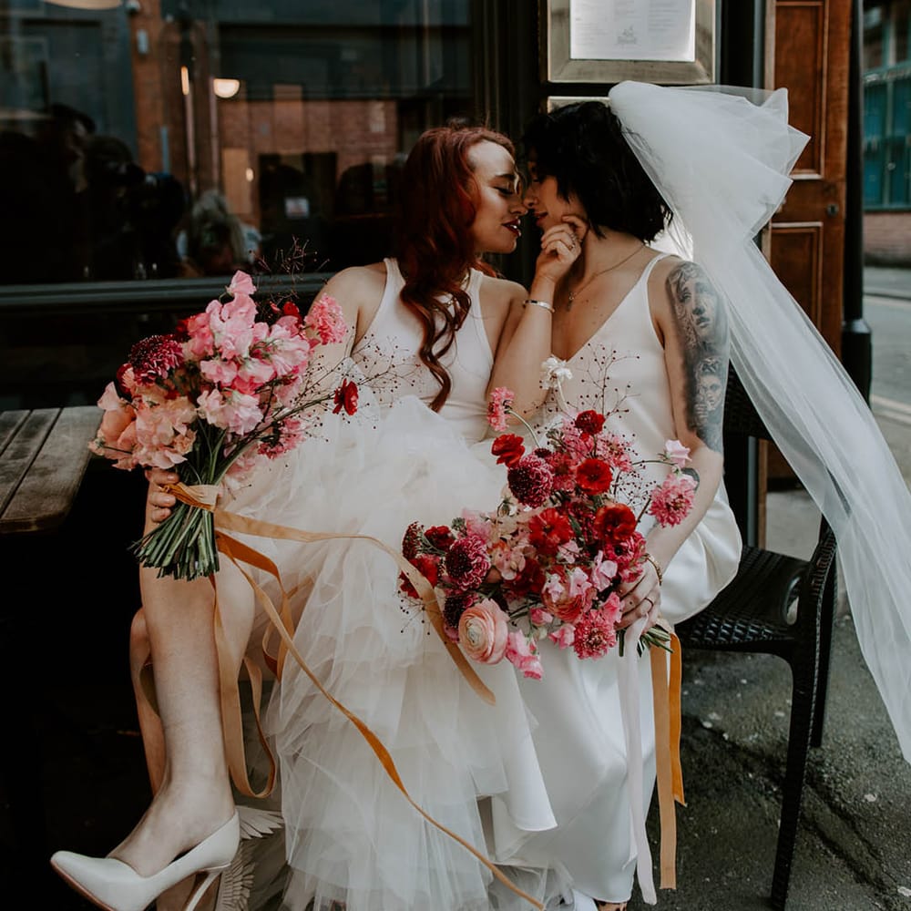 Two brides sitting and kissing in a satin slip wedding dress with big bow tulle veil and high low layered tulle wedding dress