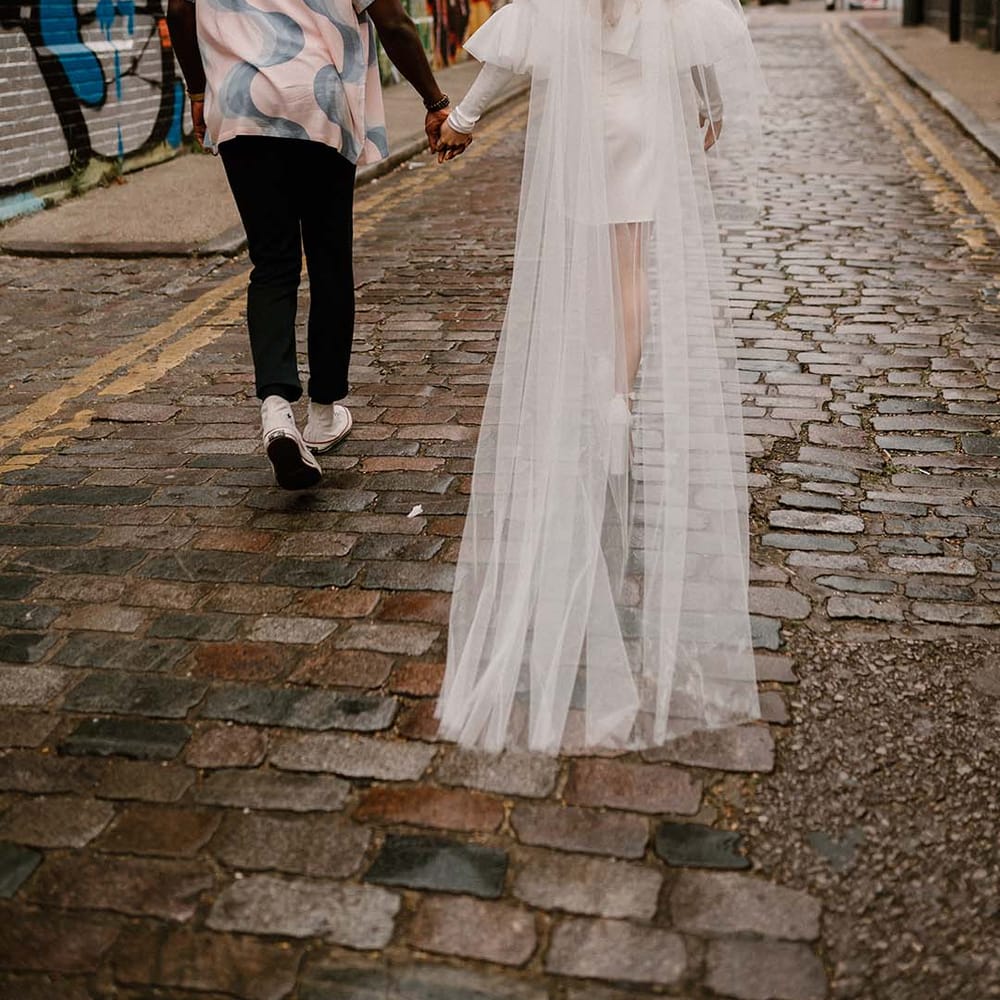 Groom in a patterned shirt and Converse trainers holding hands with his bride in a short wedding dress with long veil
