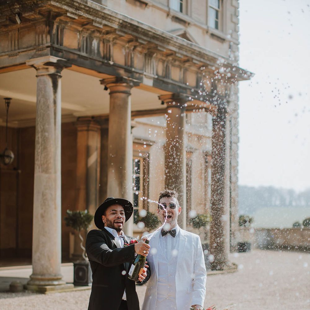Groom in a two piece black suit and fedora hat popping champagne with his partner in a white three piece groom suit, white boots and cape 