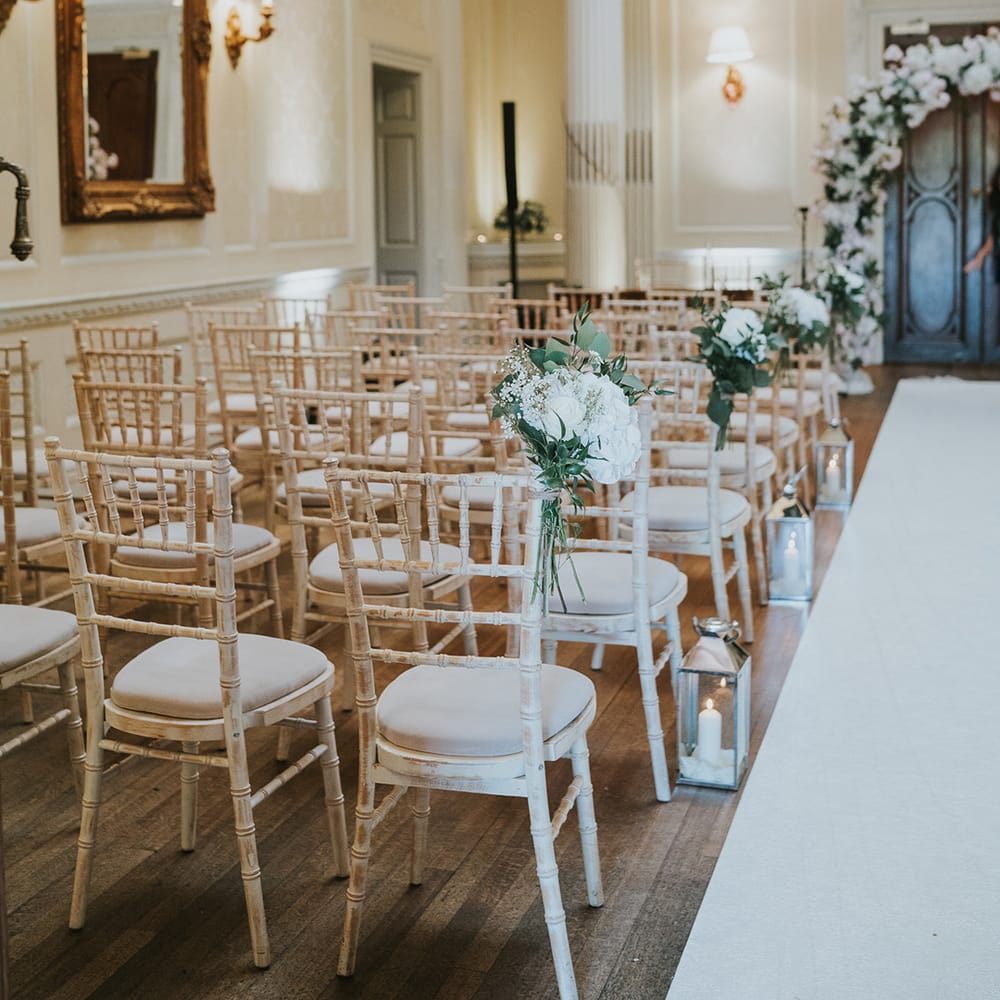 White chairs with silver candle lantern aisle decoration and white roses on the chairs  